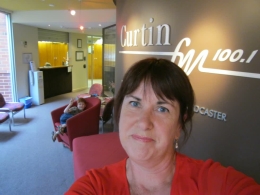 Close up of Serena in a red top in the foyer of the Curtain FM Radio station in Perth with Riley doing a peace sign in the background.